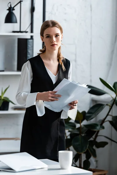 Schöne Geschäftsfrau in formeller Kleidung hält Dokumente in der Hand und blickt am Arbeitsplatz in die Kamera — Stockfoto