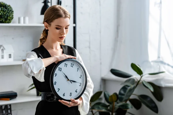 Attractive woman in formal wear holding clock at workplace — Stock Photo