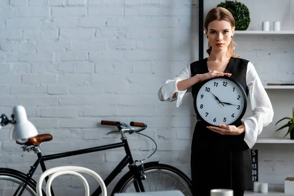 Femme en tenue formelle regardant la caméra et tenant horloge sur le lieu de travail — Photo de stock