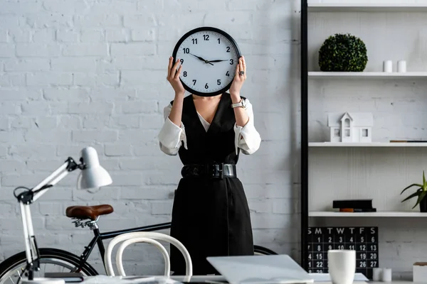 Businesswoman in formal wear holding clock in front of face in office — Stock Photo