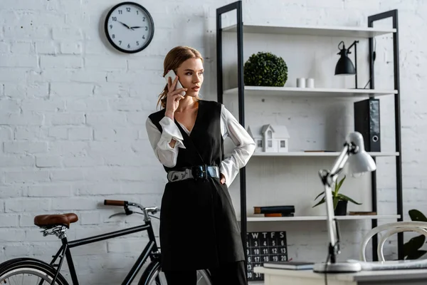 Femme d'affaires en tenue formelle avec la main sur la hanche parler sur smartphone au bureau — Photo de stock