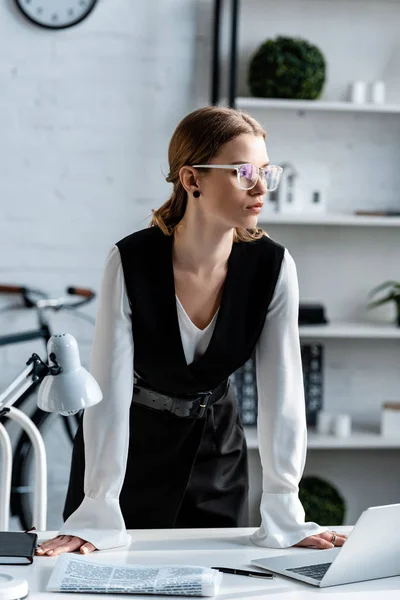 Mujer de negocios en ropa formal y gafas de pie cerca del escritorio con ordenador portátil y periódico en el lugar de trabajo - foto de stock