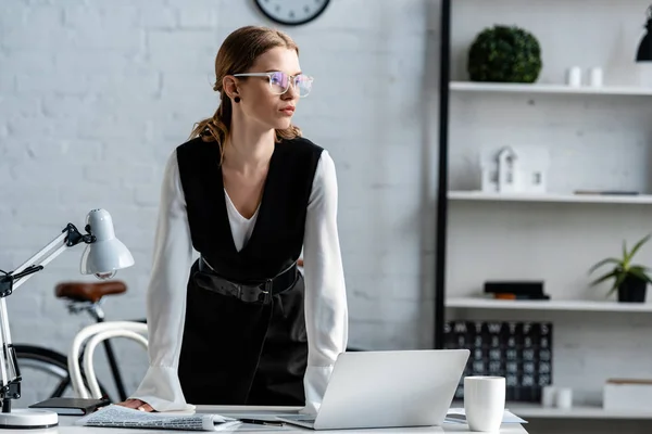 Donna d'affari in abbigliamento formale e occhiali in piedi vicino alla scrivania del computer sul posto di lavoro e guardando altrove — Foto stock