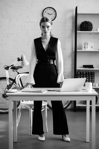 Black and white photo of businesswoman in formal wear looking at camera and standing near computer desk at workplace — Stock Photo