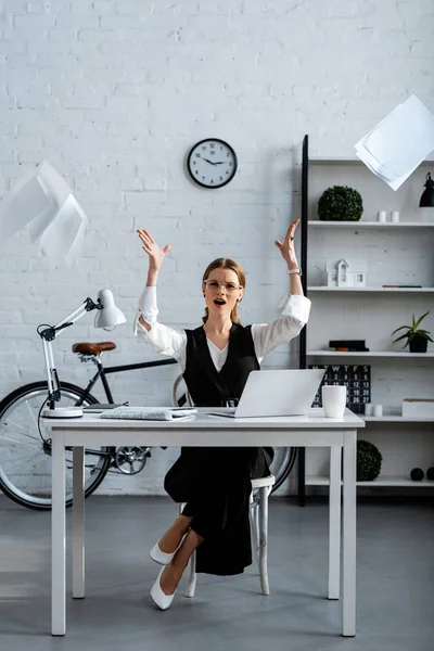Незадоволена бізнес-леді в офіційному одязі, сидячи за столом, жестикулюючи руками і кидаючи документи в повітря на робочому місці — стокове фото