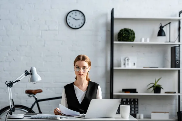 Серьезная деловая женщина в формальной одежде сидит за компьютерным столом с документами на рабочем месте — стоковое фото
