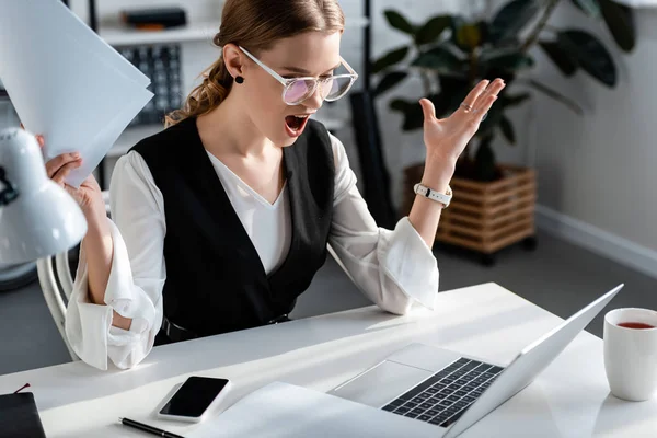 Mujer de negocios sorprendida en ropa formal sentada en el escritorio de la computadora y gesticulando con las manos en el lugar de trabajo - foto de stock