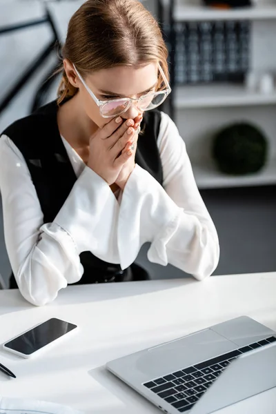 Foco seletivo de empresária chocada em desgaste formal com as mãos dobradas sentadas na mesa do computador no local de trabalho — Fotografia de Stock