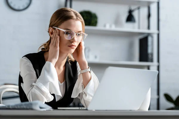 Mujer de negocios en ropa formal y gafas sentada en el escritorio de la computadora y con dolor de cabeza en el lugar de trabajo - foto de stock