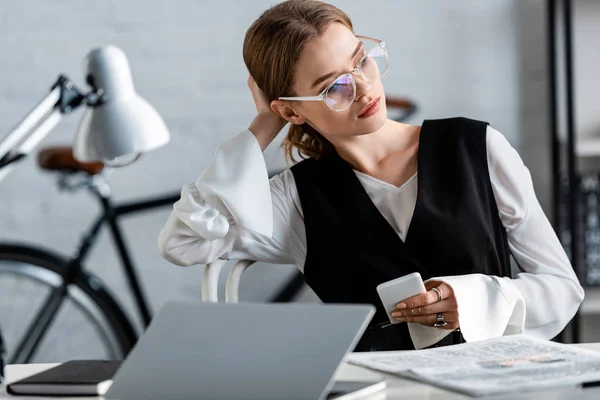 Empresária em uso formal e óculos sentados na mesa do computador e usando smartphone no local de trabalho — Fotografia de Stock