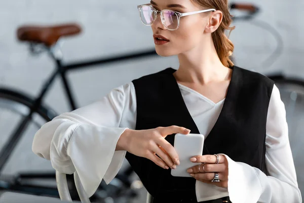 Beautiful businesswoman in formal wear and glasses using smartphone at workplace — Stock Photo