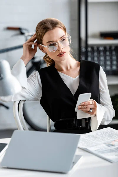 Избирательный фокус деловой женщины в формальной одежде и очках, сидящей за компьютерным столом и использующей смартфон на рабочем месте — стоковое фото