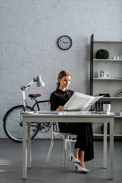 Businesswoman in black formal wear sitting at desk and reading newspaper in office — Stock Photo