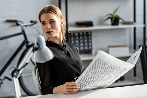 Mulher de negócios atraente em preto formal desgaste sentado na mesa e ler jornal no local de trabalho — Fotografia de Stock