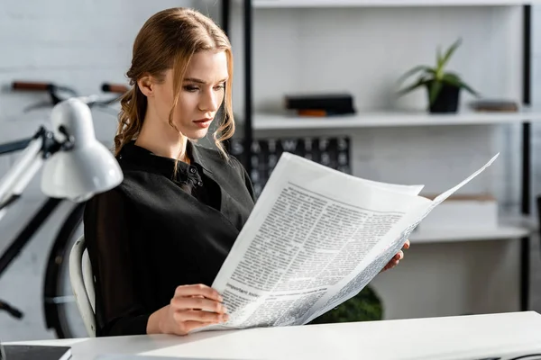 Empresaria enfocada en ropa formal negra sentada en el escritorio y leyendo el periódico en el lugar de trabajo - foto de stock