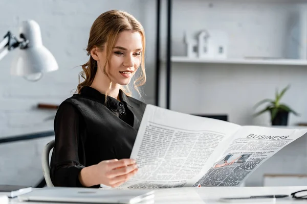 Sorridente empresária em preto formal desgaste sentado na mesa e ler jornal no local de trabalho — Fotografia de Stock