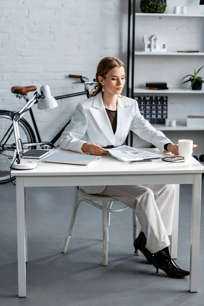 Femme d'affaires en tenue de cérémonie assis au bureau de l'ordinateur avec journal et tasse de café au bureau — Photo de stock