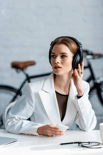 Selective focus of businesswoman in headphones sitting at desk and using smartphone at workplace — Stock Photo