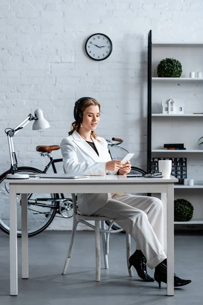 Businesswoman in headphones sitting at desk and using smartphone in office — Stock Photo