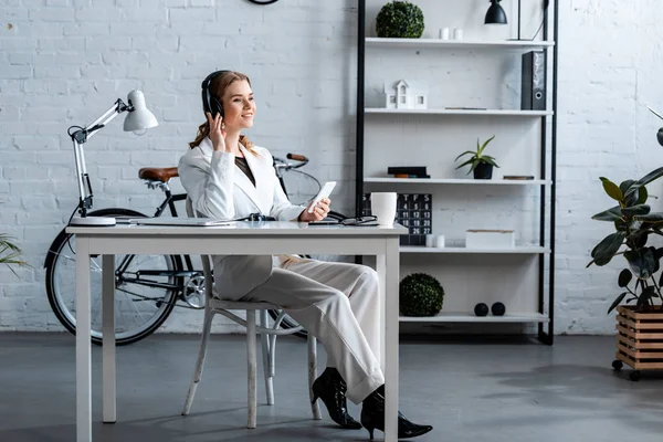 Mujer de negocios sonriente en auriculares sentados en el escritorio y utilizando el teléfono inteligente en la oficina - foto de stock