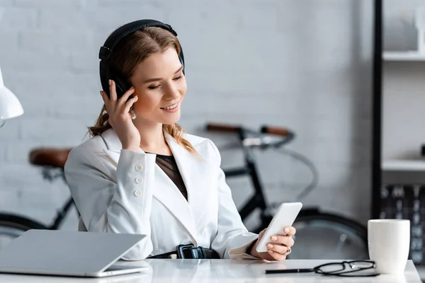 Belle femme d'affaires souriante dans les écouteurs assis au bureau et en utilisant smartphone sur le lieu de travail — Photo de stock