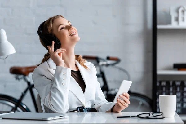 Сміється бізнес-леді в навушниках, сидячи за столом і використовуючи смартфон на робочому місці — стокове фото
