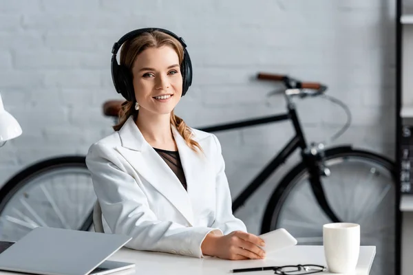 Businesswoman in headphones sitting at desk, looking at camera and holding smartphone at workplace — Stock Photo