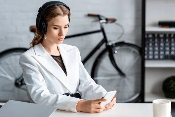 Mulher de negócios focada em fones de ouvido sentados na mesa e usando smartphone no local de trabalho — Fotografia de Stock