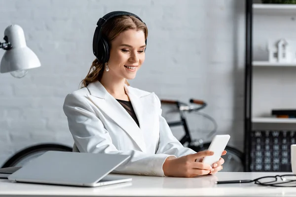 Smiling businesswoman in headphones and formal wear sitting at desk and using smartphone at workplace — Stock Photo