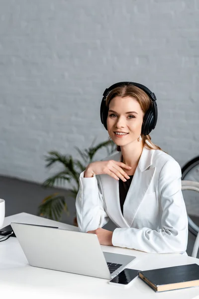 Femme d'affaires souriante dans les écouteurs assis au bureau de l'ordinateur sur le lieu de travail — Photo de stock