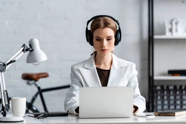 Femme d'affaires sérieuse dans les écouteurs assis au bureau de l'ordinateur sur le lieu de travail — Photo de stock