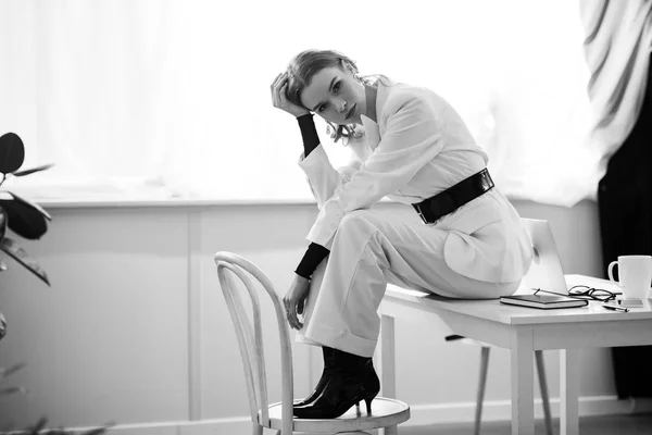 Чорно-біла фотографія стильної бізнес-леді в офіційному одязі, сидячи на столі і дивлячись на камеру на робочому місці — стокове фото