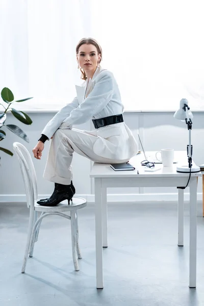 Stylish businesswoman in formal wear sitting on desk and looking at camera at workplace — Stock Photo