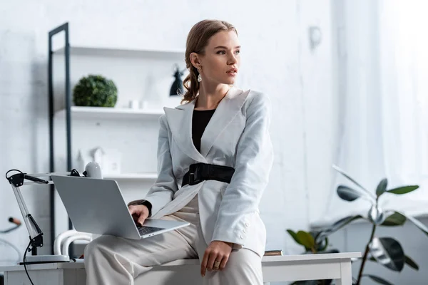 Стильная деловая женщина в белой формальной одежде сидит на столе с ноутбуком на рабочем месте и смотрит в сторону — стоковое фото