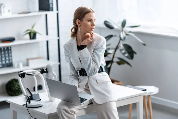 Bela mulher de negócios em branco formal desgaste sentado na mesa com laptop no local de trabalho e olhando para longe — Fotografia de Stock