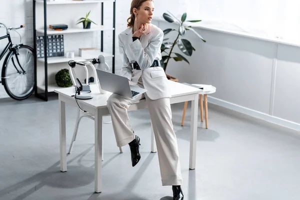Femme d'affaires en blanc vêtements formels assis sur le bureau avec ordinateur portable sur le lieu de travail et regarder ailleurs — Photo de stock