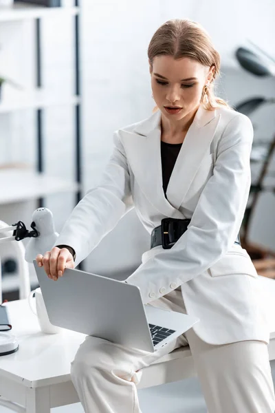 Beautiful focused businesswoman in white formal wear sitting on desk and using laptop at workplace — Stock Photo