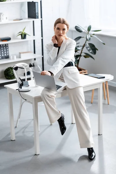 Geschäftsfrau in weißer Festkleidung sitzt am Schreibtisch mit Laptop am Arbeitsplatz und blickt in die Kamera — Stockfoto