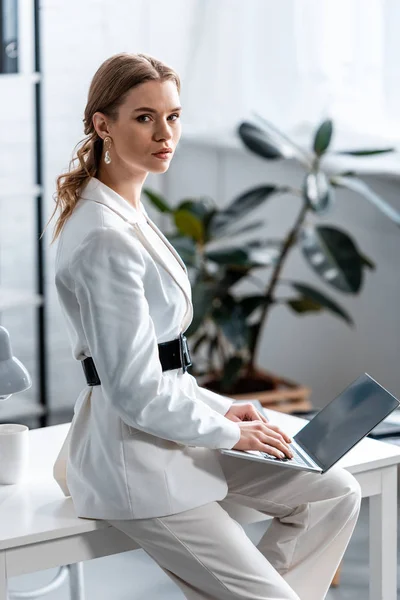 Schöne Geschäftsfrau in weißer Festkleidung sitzt am Schreibtisch, schaut in die Kamera und benutzt Laptop am Arbeitsplatz — Stockfoto