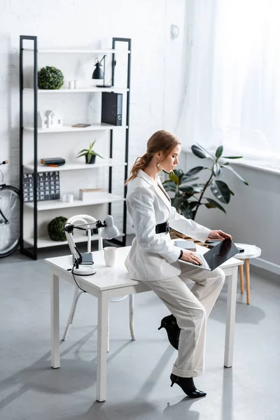 Focused businesswoman in white formal wear sitting on desk and using laptop at workplace — Stock Photo