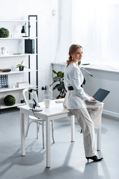 Деловая женщина в белой формальной одежде сидит на столе и использует ноутбук в офисе — стоковое фото