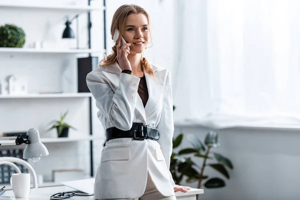 Mulher de negócios sorridente em branco formal desgaste falando no smartphone no local de trabalho — Fotografia de Stock