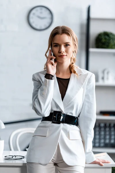 Schöne Geschäftsfrau in weißer Festkleidung, die am Arbeitsplatz mit dem Smartphone spricht und in die Kamera schaut — Stockfoto