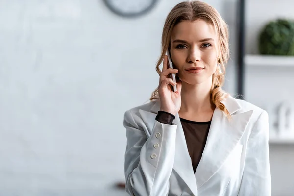 Mujer de negocios en ropa blanca formal hablando en el teléfono inteligente y mirando a la cámara en el lugar de trabajo - foto de stock