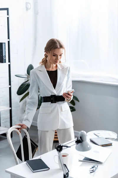 Fokussierte Geschäftsfrau in weißer Dienstkleidung mit Smartphone am Arbeitsplatz — Stockfoto
