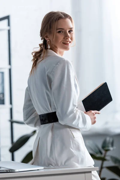 Lächelnde Geschäftsfrau in offizieller Kleidung mit Notizbuch und Blick in die Kamera am Arbeitsplatz — Stockfoto