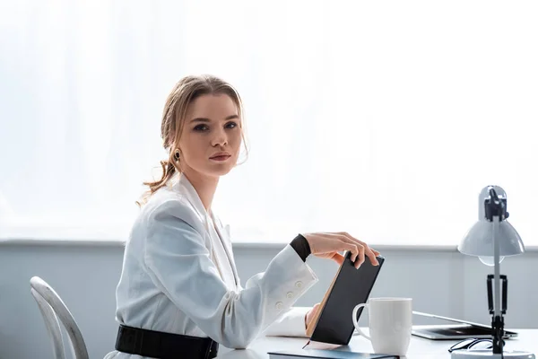 Schöne Geschäftsfrau in formeller Kleidung, die am Schreibtisch mit Notizbuch sitzt und am Arbeitsplatz in die Kamera schaut — Stockfoto