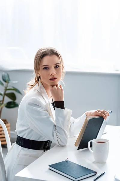 Hermosa mujer de negocios sentada en el escritorio, sosteniendo el cuaderno y mirando la cámara en el lugar de trabajo - foto de stock
