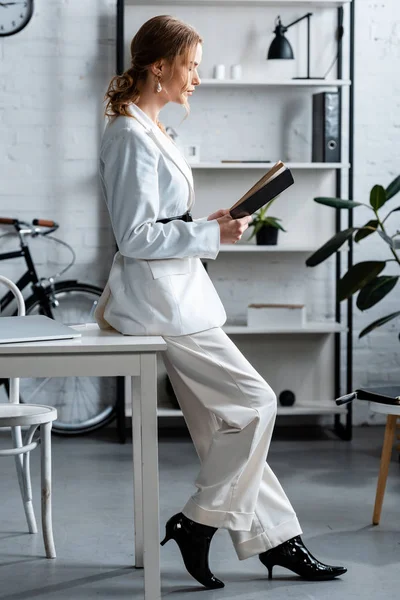 Stylish businesswoman in formal wear sitting on desk and reading notebook at workplace — Stock Photo