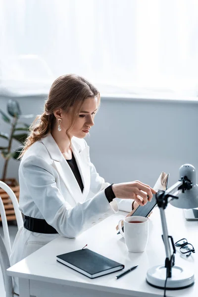 Focused businesswoman in formal wear sitting at desk and reading notebook at workplace — Stock Photo